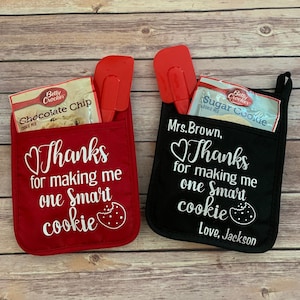 May include: Two red and black oven mitts with a pocket for a spatula and a package of baking mix. The mitts have the text "Thanks for making me one smart cookie" with a heart and a cookie graphic. One mitt has the name "Mrs. Brown" and the other has the name "Love, Jackson".