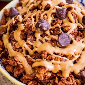 May include: A close-up of a bowl of chocolate granola with peanut butter drizzled on top and chocolate chips scattered throughout.