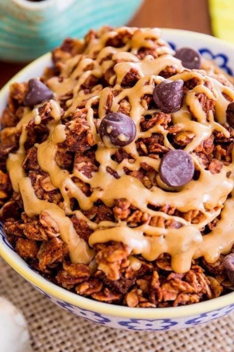 May include: A close-up of a bowl of chocolate granola with peanut butter drizzled on top and chocolate chips scattered throughout.