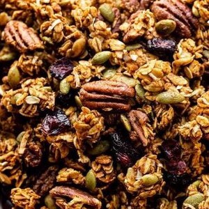 May include: A close-up of a bowl of granola with pecans, pumpkin seeds, and dried cranberries. The granola is a light brown color and the pecans are a darker brown. The pumpkin seeds are green and the cranberries are red.