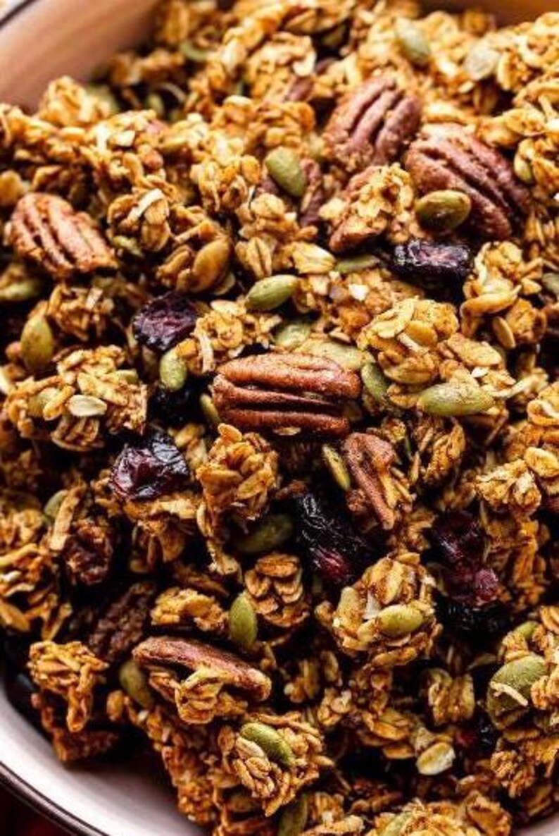 May include: A close-up of a bowl of granola with pecans, pumpkin seeds, and dried cranberries. The granola is a light brown color and the pecans are a darker brown. The pumpkin seeds are green and the cranberries are red.