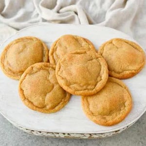 May include: A close-up of six golden brown cookies on a white oval plate. The cookies are round and have a slightly crispy texture.