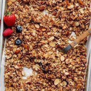 May include: A close-up of a baking sheet filled with homemade granola. The granola is a mixture of oats, almonds, and other ingredients. The granola is golden brown and looks crispy.