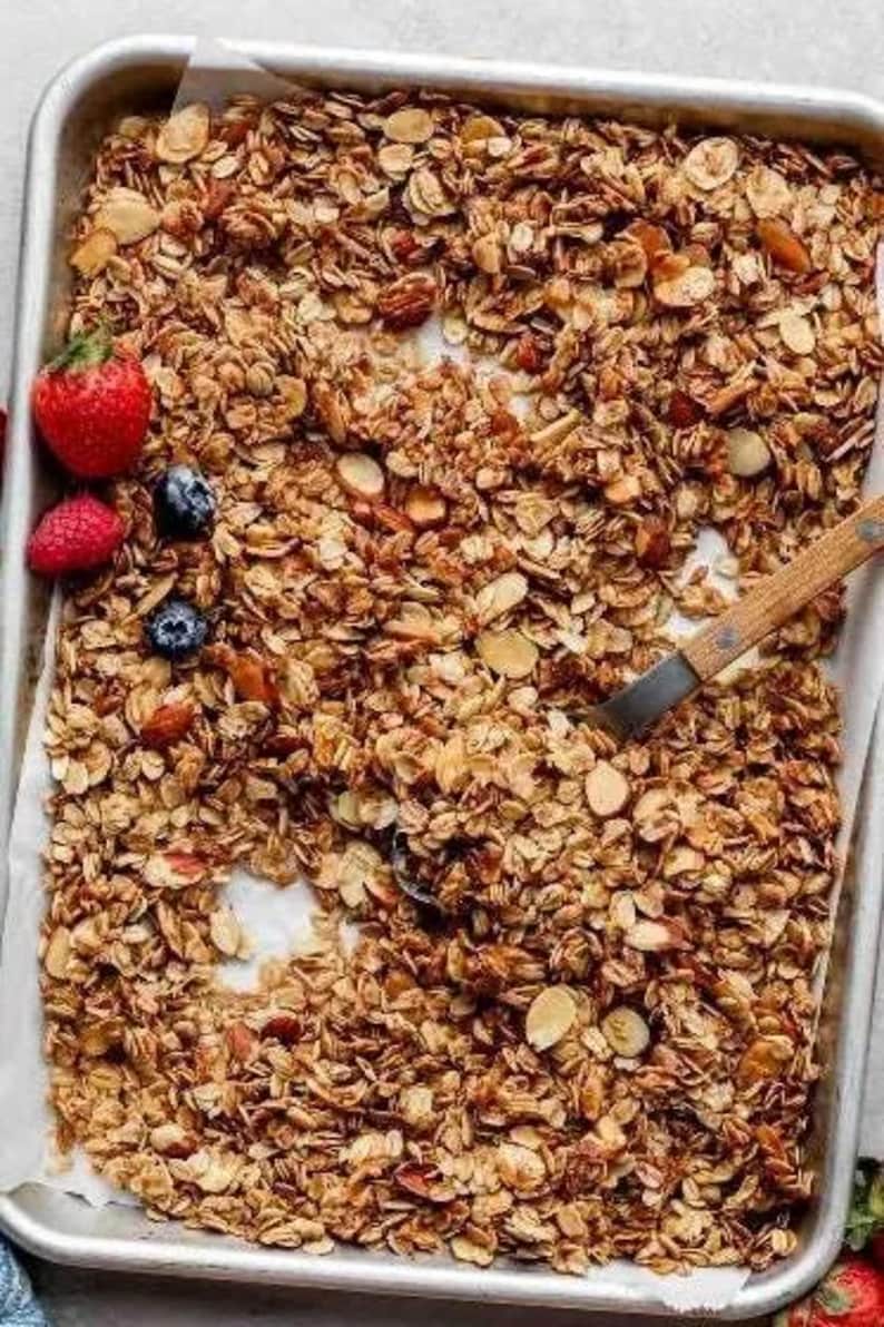 May include: A close-up of a baking sheet filled with homemade granola. The granola is a mixture of oats, almonds, and other ingredients. The granola is golden brown and looks crispy.