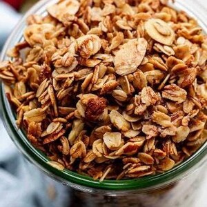 May include: A close-up of a glass jar filled with granola. The granola is a mixture of oats, nuts, and seeds. It is a light brown color and has a slightly crunchy texture.
