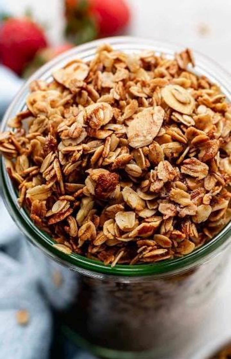 May include: A close-up of a glass jar filled with granola. The granola is a mixture of oats, nuts, and seeds. It is a light brown color and has a slightly crunchy texture.