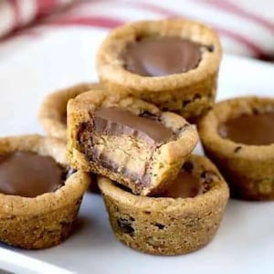May include: A close-up of several peanut butter cup cookies on a white plate. The cookies have a chocolate center and are surrounded by a golden-brown cookie base. One cookie is cut in half, revealing the peanut butter filling.