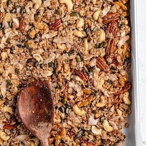May include: A baking sheet filled with homemade granola with a wooden spoon. The granola is made with oats, nuts, seeds, and coconut flakes.