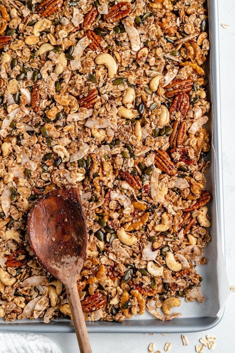 May include: A baking sheet filled with homemade granola with a wooden spoon. The granola is made with oats, nuts, seeds, and coconut flakes.