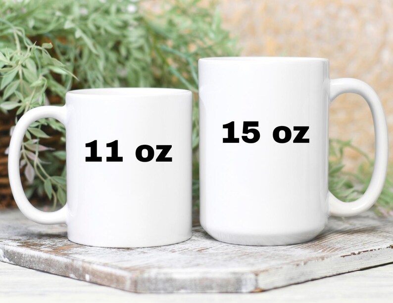 Op de afbeelding: Twee witte keramische mokken met zwarte tekst. De kleinere mok is 11 oz en de grotere mok is 15 oz.