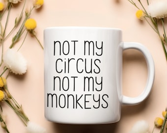 Op de afbeelding: Witte keramische mok met zwarte tekst die "not my circus not my monkeys" luidt, omringd door witte en gele bloemen op een lichtroze achtergrond.