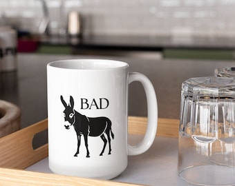 Op de afbeelding: Witte keramische koffiemok met een zwart silhouet van een ezel en het woord "BAD" erop gedrukt.