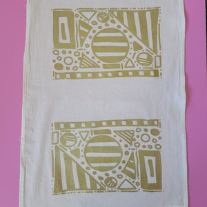 May include: A white tea towel with two gold geometric designs. The designs feature circles, squares, triangles, and stripes.