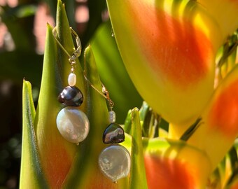 May include: Gold drop earrings with white and dark grey baroque pearls. The earrings are suspended from a small gold hoop. The background features yellow, orange, and green tropical foliage.