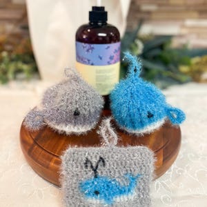 May include: Three crocheted whale-shaped bath poufs in gray and blue. The poufs are on a wooden surface. A bottle of liquid soap is in the background.