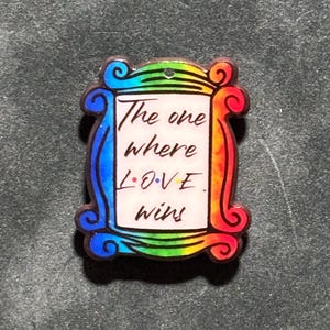 May include: A rectangular acrylic pendant featuring a rainbow-colored frame resembling a picture frame. The center is white with black text that reads The one where L.O.V.E. wins, with colored dots between the letters of the word LOVE.