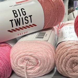 May include: Assortment of Big Twist Value yarn skeins in various shades of pink and peach. The yarn is 100% acrylic. The label on the top skein reads "BIG TWIST VALUE". The yarn is tightly wound into balls, ready for knitting or crocheting projects.