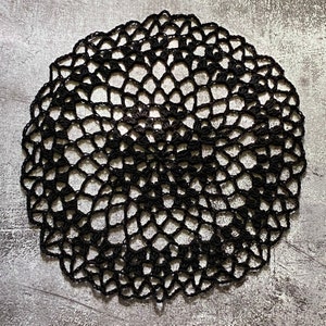 May include: A circular black crochet doily featuring an intricate, openwork web-like pattern. The design radiates from the center with repeating loops and small clusters of stitches, creating a delicate, lacy texture throughout the entire piece.