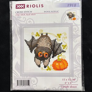 May include: A Riolis 1918 cross-stitch kit featuring a brown bat hanging upside down with orange eyes next to an orange pumpkin and yellow vines. The packaging includes the text Riolis, 1918, Cross-Stitch, Wool/Acrylic, and 6" x 6".