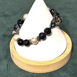 May include: A black and white gemstone bracelet with white pearl accents and a flower charm. The bracelet is displayed on a white cone-shaped stand.