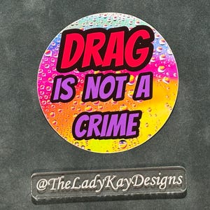 May include: A round sticker with the text "DRAG IS NOT A CRIME" in red and purple, set against a colorful, rainbow-gradient background. Below the sticker is a clear rectangular label with the text "@TheLadyKayDesigns".