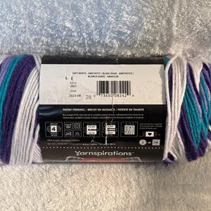 Pode incluir: Um novelo de fio acr&iacute;lico de peso m&eacute;dio 4 com um padr&atilde;o de cores variadas em branco, roxo e azul-petr&oacute;leo. O r&oacute;tulo do produto exibe o texto Yarnspirations, Soft White, Amethyst, e inclui instru&ccedil;&otilde;es de cuidado e um c&oacute;digo de barras.