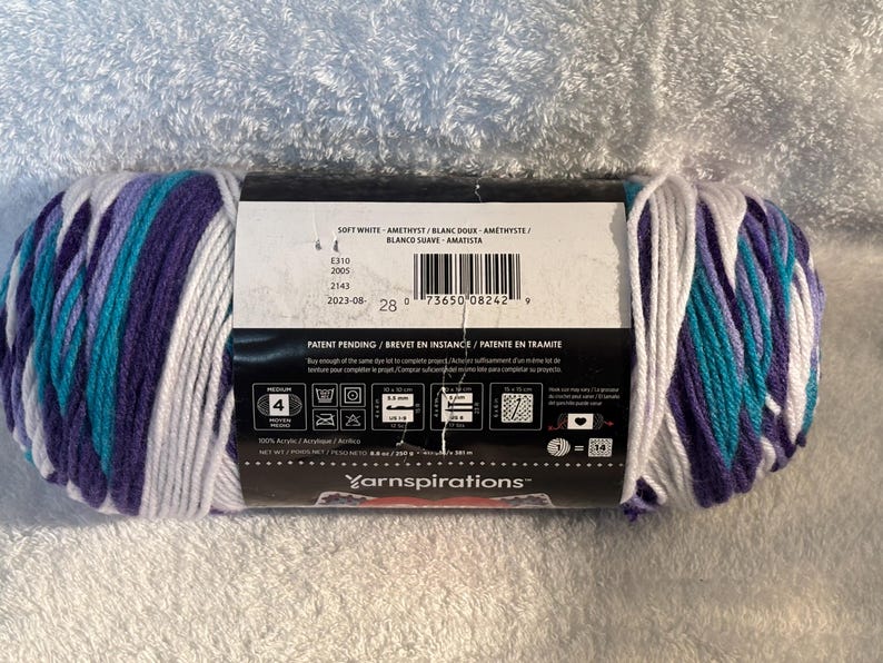Pode incluir: Um novelo de fio acr&iacute;lico de peso m&eacute;dio 4 com um padr&atilde;o de cores variadas em branco, roxo e azul-petr&oacute;leo. O r&oacute;tulo do produto exibe o texto Yarnspirations, Soft White, Amethyst, e inclui instru&ccedil;&otilde;es de cuidado e um c&oacute;digo de barras.