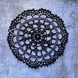 May include: A circular crochet doily featuring a complex, openwork pattern. The design is primarily black with a single concentric ring of dark gray yarn integrated into the center. The edges are finished with a scalloped, decorative border.
