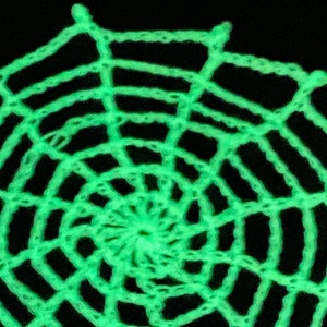 May include: A bright neon green crocheted spider web pattern featuring a circular design with radiating spokes and concentric rings. The yarn texture is clearly visible against a solid black background, creating a high-contrast visual effect.