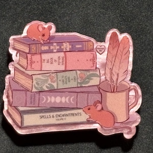 May include: A sticker featuring a stack of books with titles like "Spells & Enchantments" and "The Big Book of Potion Making." Two small orange mice are perched on the books. A mug with feathers is next to the books. The sticker has a pink and purple color scheme.