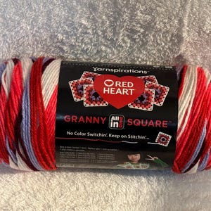 Pode incluir: Um novelo de fio Red Heart Granny All in One Square com uma paleta de cores variadas em vermelho, branco e azul claro. O r&oacute;tulo preto do produto exibe o log&oacute;tipo da marca, texto e um gr&aacute;fico de quadrados granny de croch&eacute;.