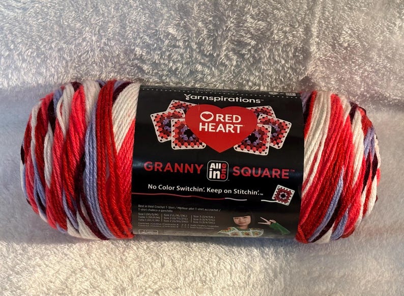 Pode incluir: Um novelo de fio Red Heart Granny All in One Square com uma paleta de cores variadas em vermelho, branco e azul claro. O r&oacute;tulo preto do produto exibe o log&oacute;tipo da marca, texto e um gr&aacute;fico de quadrados granny de croch&eacute;.