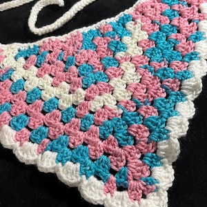 May include: A triangular crochet bandana featuring a granny square pattern in alternating colors of light pink, bright turquoise, and off-white yarn. The edges are finished with a scalloped white border and include long crocheted ties at the top.