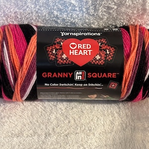 Pode incluir: Um novelo de fio Red Heart Granny All in One Square com uma paleta de cores variadas em preto, rosa brilhante, laranja e branco. O r&oacute;tulo de papel preto exibe o log&oacute;tipo da Yarnspirations, um cora&ccedil;&atilde;o vermelho e o texto Granny All in One Square.
