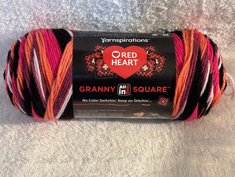 Pode incluir: Um novelo de fio Red Heart Granny All in One Square com uma paleta de cores variadas em preto, rosa brilhante, laranja e branco. O r&oacute;tulo de papel preto exibe o log&oacute;tipo da Yarnspirations, um cora&ccedil;&atilde;o vermelho e o texto Granny All in One Square.