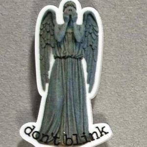 May include: A figurine of a weeping angel with gray wings and a draped gown. The angel covers its eyes with its hands. The words "don't blink" are printed in black at the bottom of the figurine.