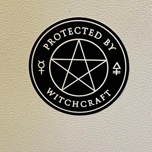May include: A circular black sticker featuring a white pentagram at the center, surrounded by the text "PROTECTED BY WITCHCRAFT" in a white serif font, with small white alchemical symbols on the left and right sides of the central star.