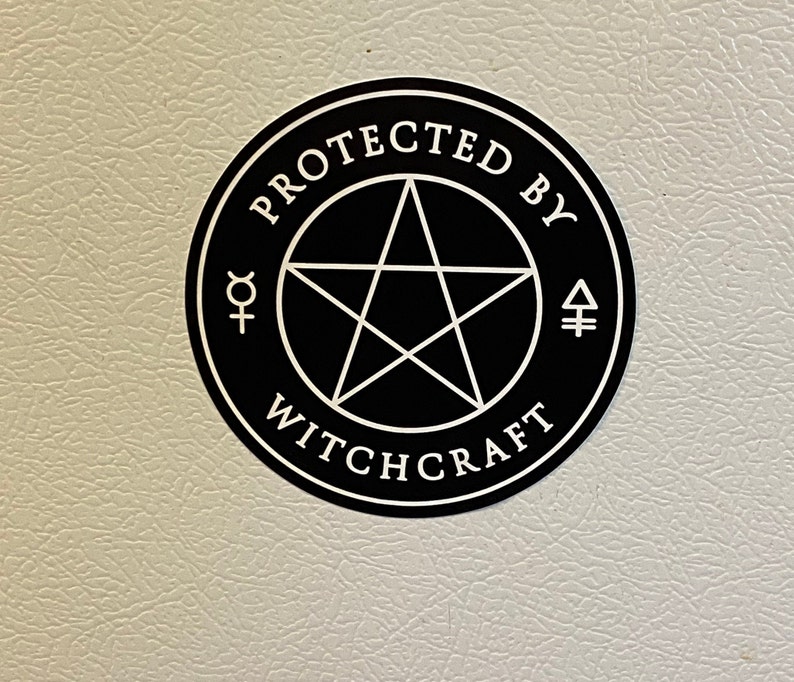 May include: A circular black sticker featuring a white pentagram at the center, surrounded by the text "PROTECTED BY WITCHCRAFT" in a white serif font, with small white alchemical symbols on the left and right sides of the central star.