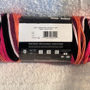 Pode incluir: Um novelo de fio acr&iacute;lico de peso m&eacute;dio 4 com um padr&atilde;o de cores variadas em preto, rosa brilhante, branco e laranja. O r&oacute;tulo central de papel exibe o texto Black-Carnation, um c&oacute;digo de barras e instru&ccedil;&otilde;es de cuidado com uma agulha de 5,0 mm.