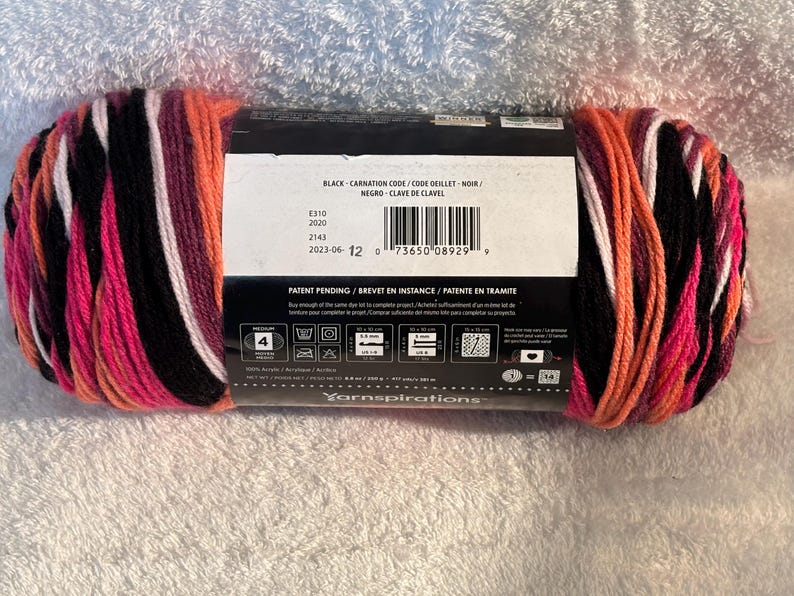 Pode incluir: Um novelo de fio acr&iacute;lico de peso m&eacute;dio 4 com um padr&atilde;o de cores variadas em preto, rosa brilhante, branco e laranja. O r&oacute;tulo central de papel exibe o texto Black-Carnation, um c&oacute;digo de barras e instru&ccedil;&otilde;es de cuidado com uma agulha de 5,0 mm.