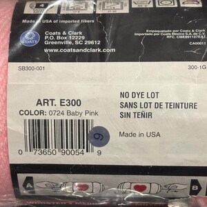 May include: A skein of baby pink yarn wrapped in a black and white paper label. The label displays the text Coats & Clark, Art. E300, Color: 0724 Baby Pink, No Dye Lot, and Made in USA, along with a barcode and various laundry care symbols.
