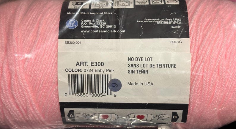 May include: A skein of baby pink yarn wrapped in a black and white paper label. The label displays the text Coats & Clark, Art. E300, Color: 0724 Baby Pink, No Dye Lot, and Made in USA, along with a barcode and various laundry care symbols.