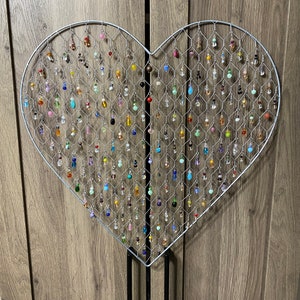 May include: A heart-shaped wire display filled with colorful beaded earrings. The earrings are various shapes and sizes, with a mix of clear, opaque, and iridescent beads. The display is mounted on a black stand, perfect for showcasing jewelry.