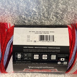 Pode incluir: Um novelo de fio multicolorido com riscas em vermelho, branco, roxo claro e bordeaux escuro. A etiqueta de papel apresenta o texto Soft White - Hot Stuff, Yarnspirations, 100% acr&iacute;lico, 250 g, 381 m, um c&oacute;digo de barras e &iacute;cones de cuidado.