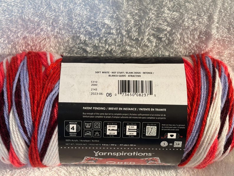 Pode incluir: Um novelo de fio multicolorido com riscas em vermelho, branco, roxo claro e bordeaux escuro. A etiqueta de papel apresenta o texto Soft White - Hot Stuff, Yarnspirations, 100% acr&iacute;lico, 250 g, 381 m, um c&oacute;digo de barras e &iacute;cones de cuidado.