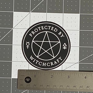 May include: A circular black sticker with a white pentagram in the center, surrounded by the text "PROTECTED BY WITCHCRAFT" and two small alchemical symbols. A metal ruler shows the sticker has a diameter of approximately 3.5 inches.