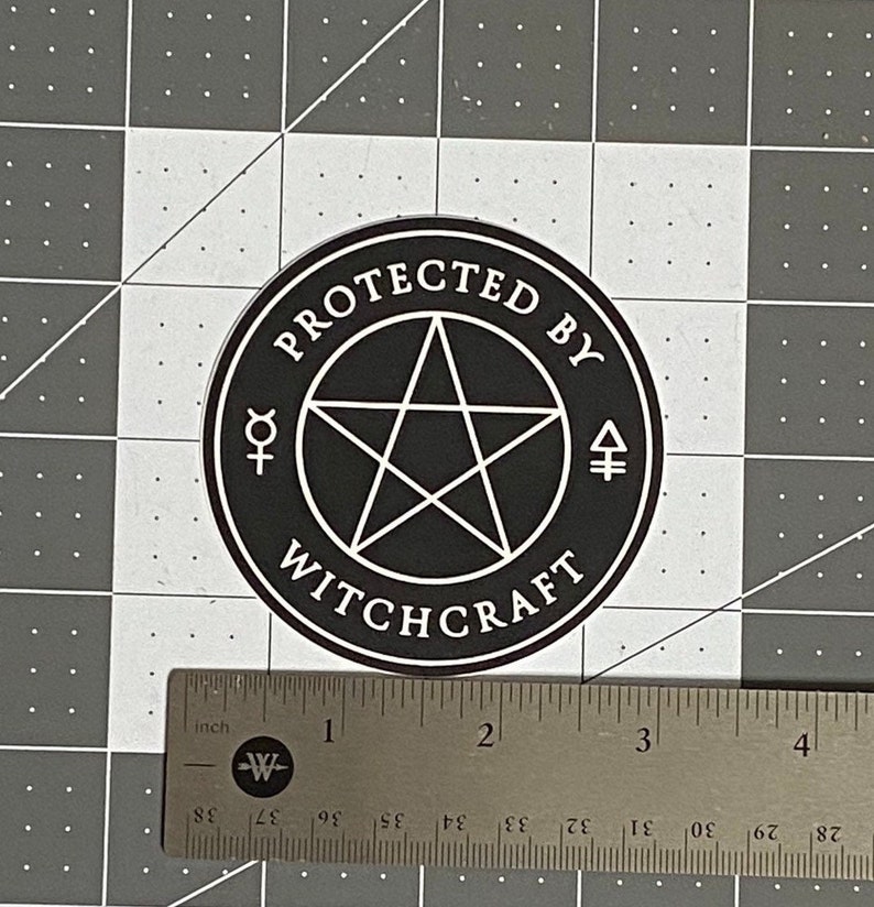 May include: A circular black sticker with a white pentagram in the center, surrounded by the text "PROTECTED BY WITCHCRAFT" and two small alchemical symbols. A metal ruler shows the sticker has a diameter of approximately 3.5 inches.