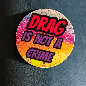 May include: A round sticker with a rainbow background and water droplets. The text "DRAG IS NOT A CRIME" is written in bold, black letters with a red outline.