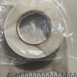 May include: A roll of white adhesive tape encased in a clear plastic bag. The inner cardboard core of the tape features the repeated blue text PRO. A ruler positioned below the tape shows a diameter of approximately 4.5 inches for the roll.