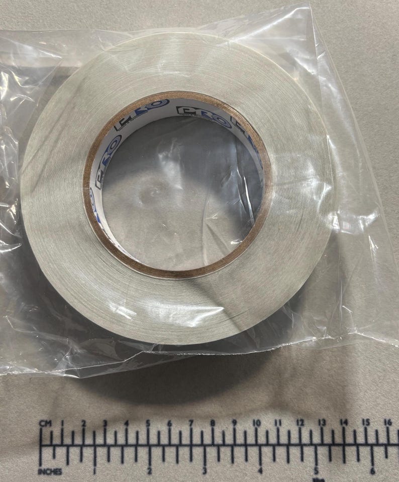 May include: A roll of white adhesive tape encased in a clear plastic bag. The inner cardboard core of the tape features the repeated blue text PRO. A ruler positioned below the tape shows a diameter of approximately 4.5 inches for the roll.